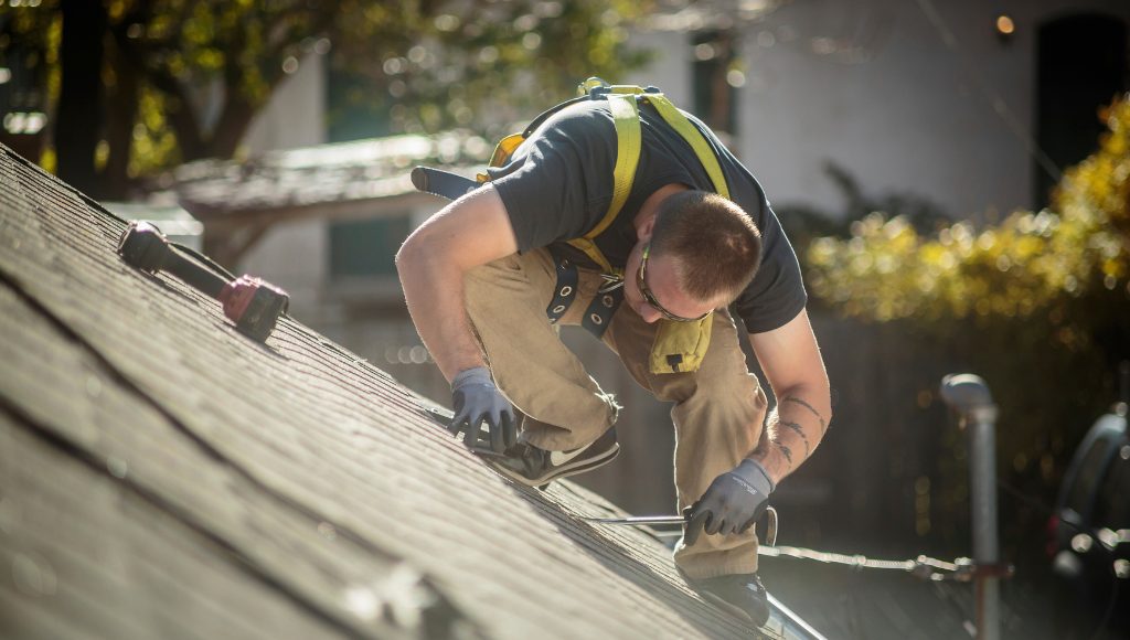 A roofer is crouched down on a residential roof repairing damaged shingles.