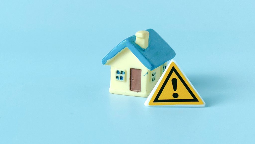 A yellow warning triangle with an exclamation point leaning against a tiny model house on a blue background.