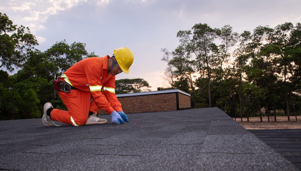 A roofer in orange coveralls with reflective arm bands, wearing a yellow hard hat works on an asphalt shingle roof.
