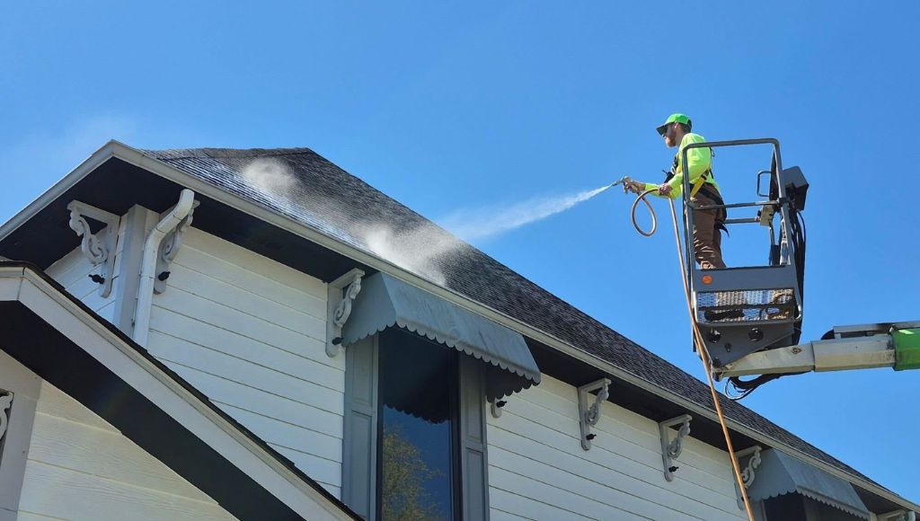 A professional roofer in a cherry picker sprays Roof Maxx rejuvenation treatment onto a steep roof.