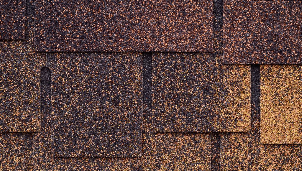 Dark brown asphalt shingles with missing granules, showing the lighter beige of the shingle tile underneath.