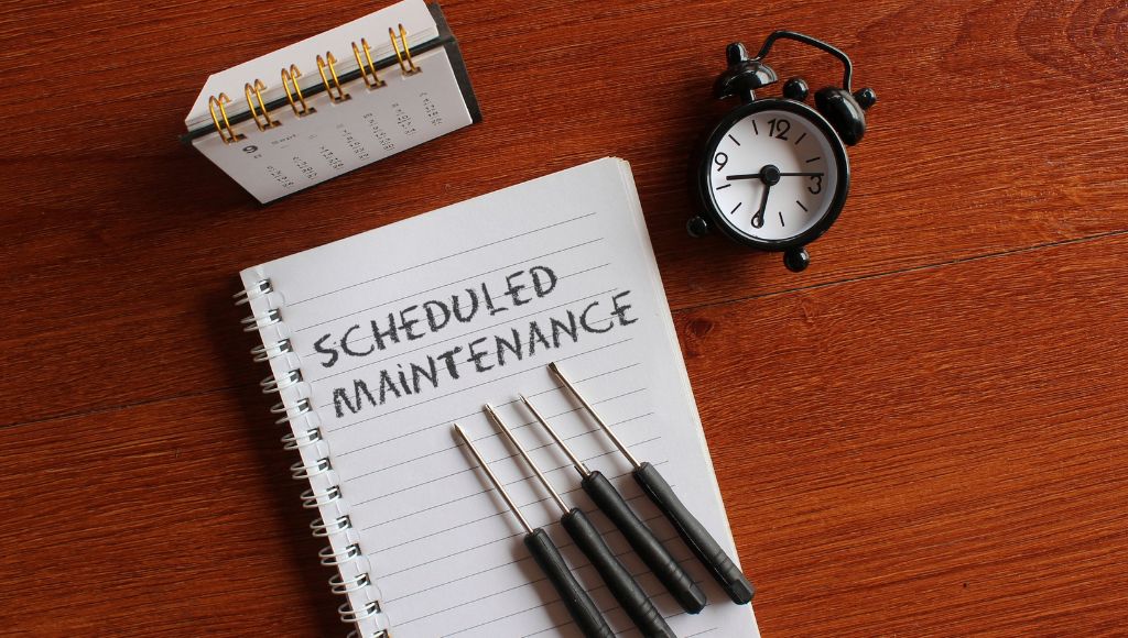A notepad with the words "SCHEDULED MAINTENANCE" is displayed with screwdrivers and an alarm clock, and a calendar on a wood-grain surface.